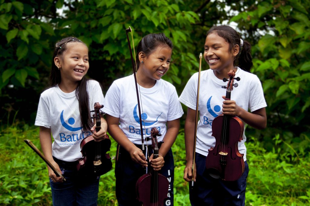 Tres niñas indígenas sonrientes sostienen sus violines en Puerto Nariño, Amazonas.
