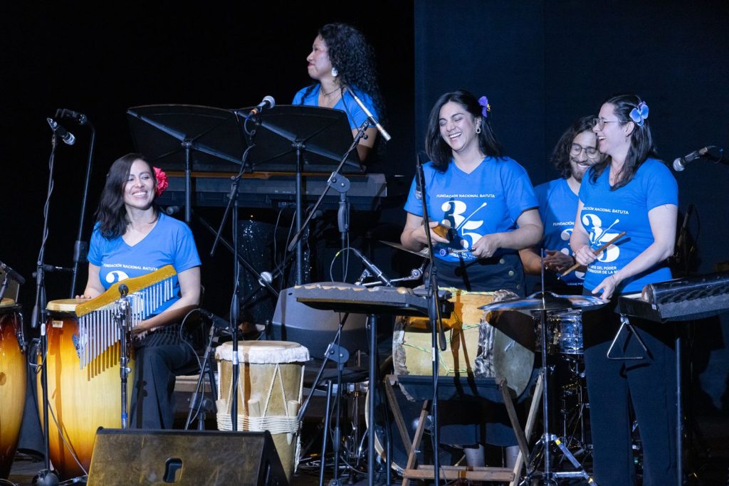 Mujeres integrantes de la orquesta Batuta sonríen durante concierto junto con instrumentos de percusión