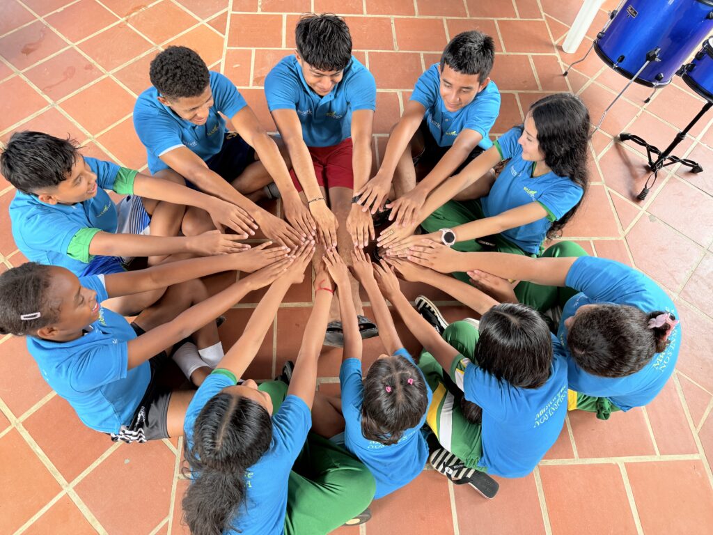Un grupo de niños y niñas sentados en  círculo en el piso de un salón con sus manos extendidas al centro