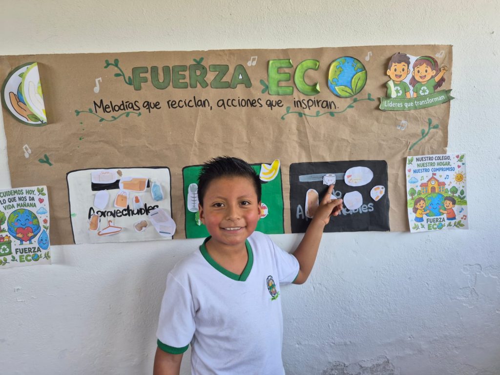 Un niño sonriendo apuntando a cartelera sobre el cuidado ambiental pegada en la pared de un salón
