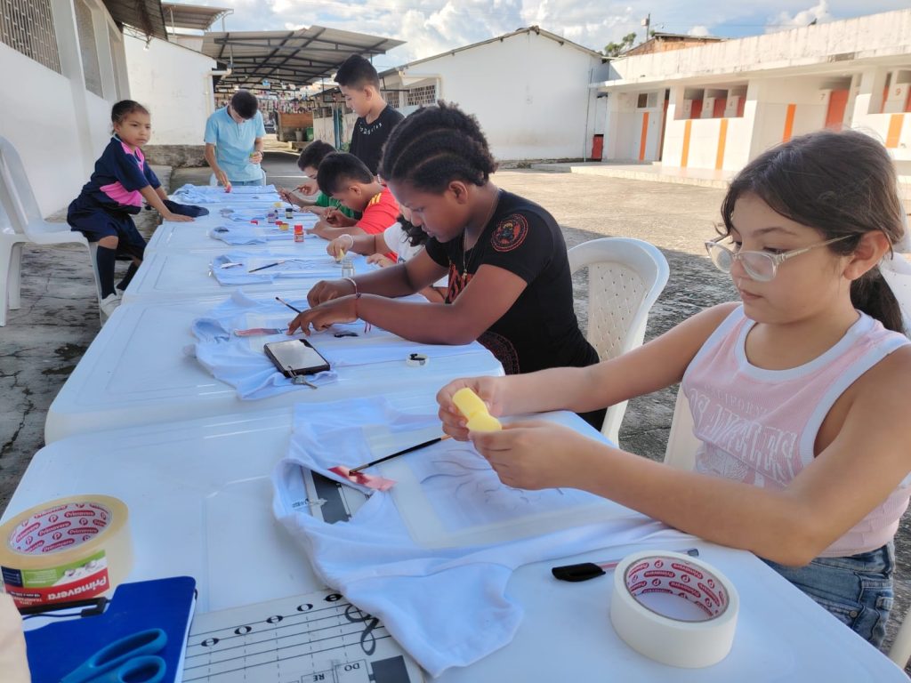 Niños y niñas sentados al aire libre realizando manualidades con camisetas  y pintura